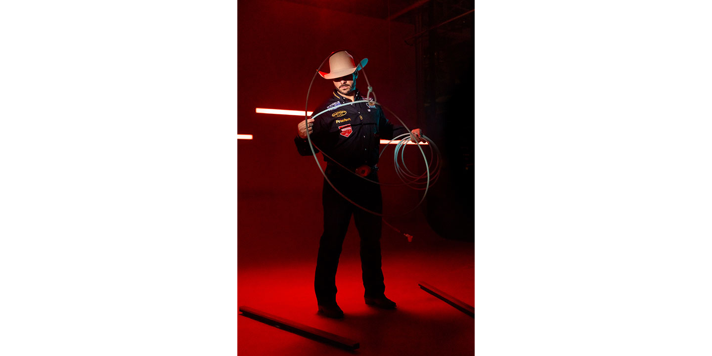 Anthony Lucia vistiendo una camisa negra y un sombrero de vaquero, sosteniendo una cuerda con iluminación roja detrás de él.